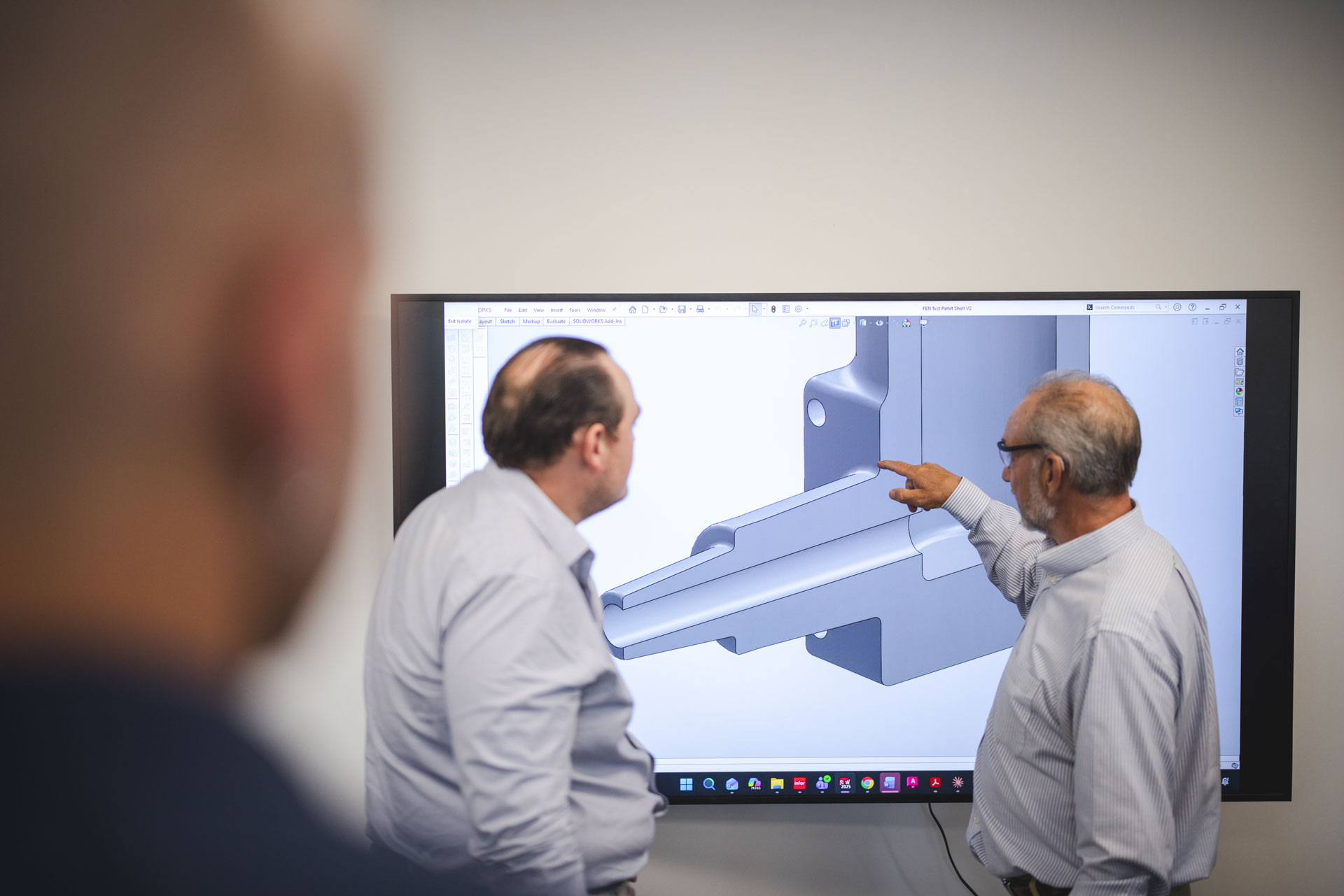 Two men in dress shirts discuss a 3D model displayed on a large screen, while a blurred person observes in the foreground. One man points at the screen, indicating a specific detail on the model.