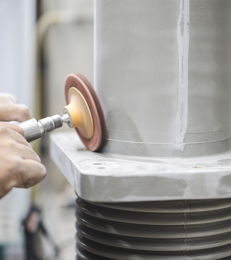 A person uses a handheld power tool with a polishing wheel to buff a large metal or ceramic cylindrical object, focusing on smoothing the surface near the base.