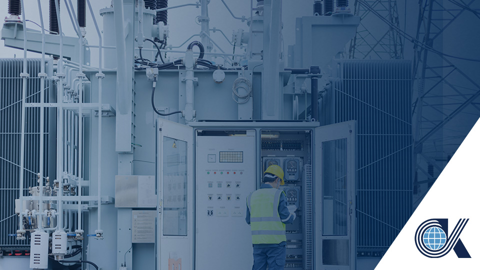 A worker in a yellow vest and helmet operates an outdoor control panel surrounded by large industrial equipment. A blue geometric logo appears in the lower right corner.