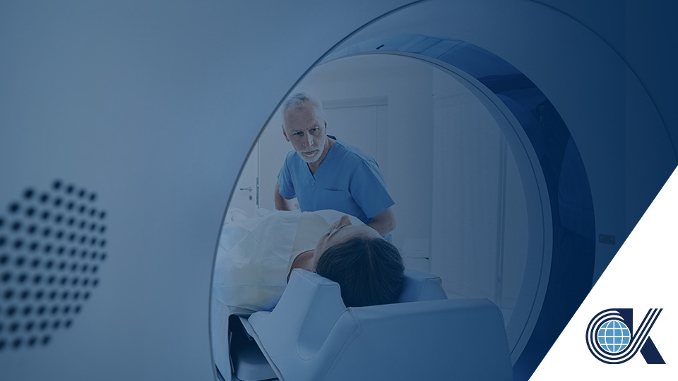 A medical professional prepares a patient for an MRI scan, with the patient lying on a table being moved into the MRI machine. The scene takes place in a clinical setting. A logo is visible in the lower right corner.