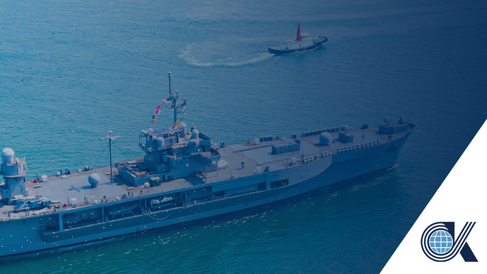 A large battleship sails in blue water, with a smaller boat nearby. The lower right corner features a stylized OK logo in white and blue.