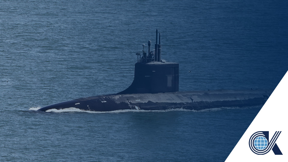 A black submarine partially surfaced in calm blue water, with an overlay and a circular logo featuring a globe and the letter K in the lower right corner.
