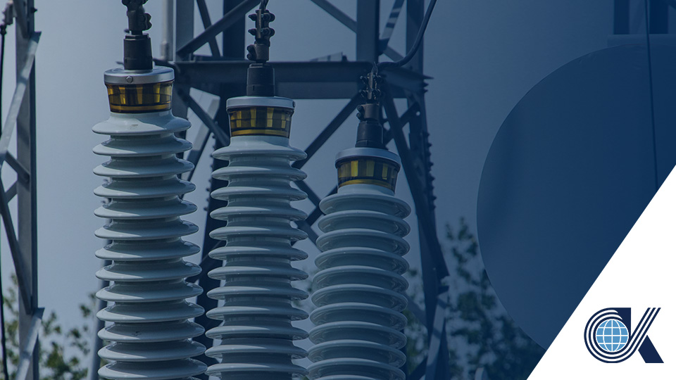 Close-up of three electrical insulators on a power line structure with a blue globe and OK logo in the lower right corner. Trees and sky are visible in the background.