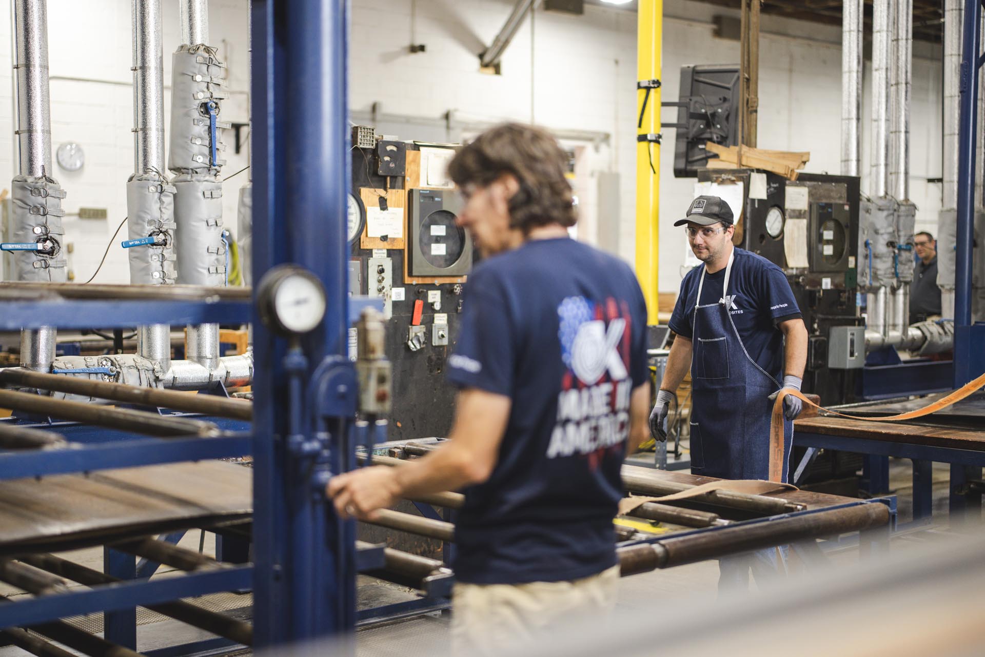 Two workers operate machinery in a factory with metal pipes and gauges. One worker wears a Made in America shirt, and the other wears a cap, both focused on their tasks in the industrial setting.