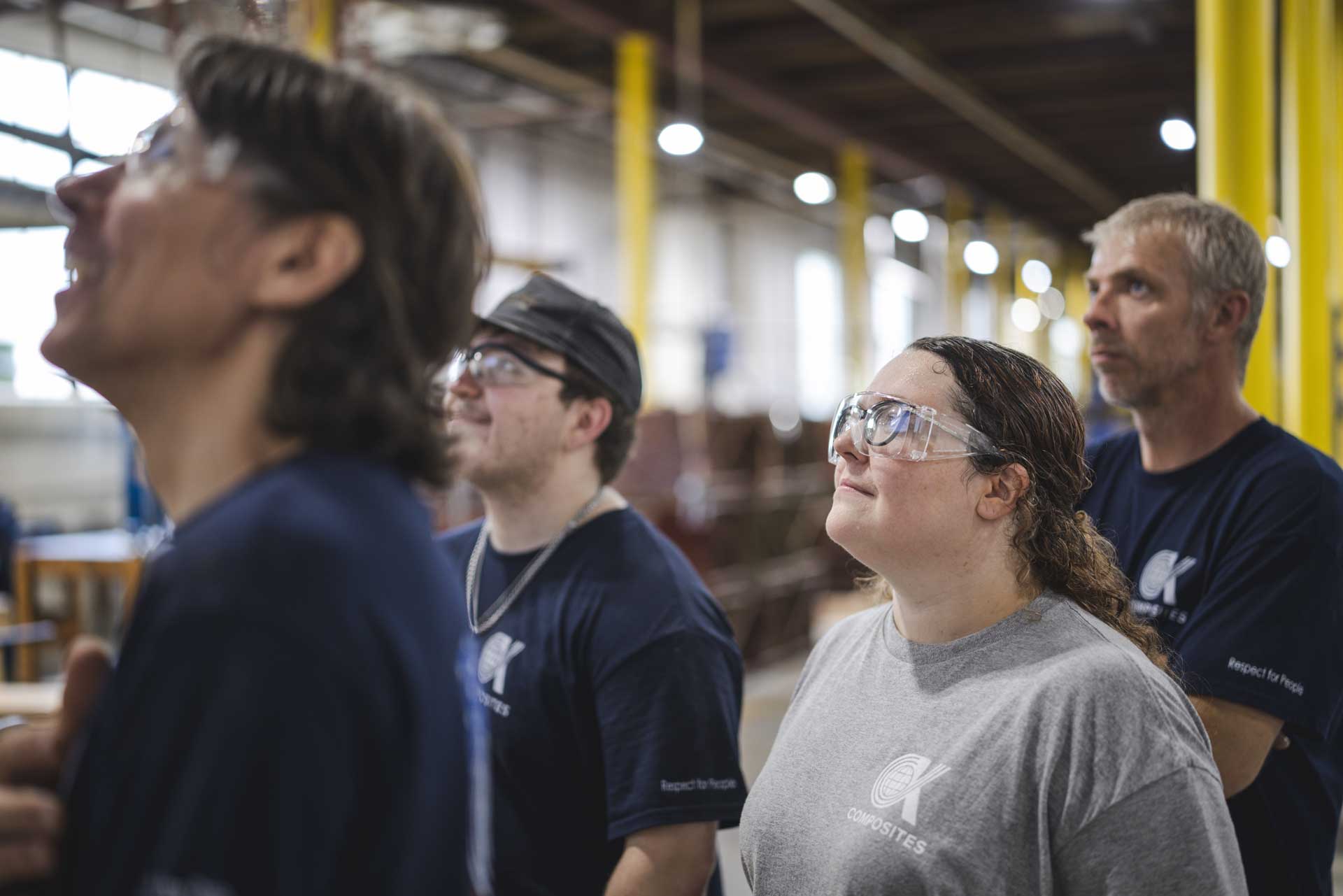 A group of four people wearing safety glasses and work shirts stand indoors, looking upward and appearing focused. The background shows an industrial or factory setting with yellow pillars.