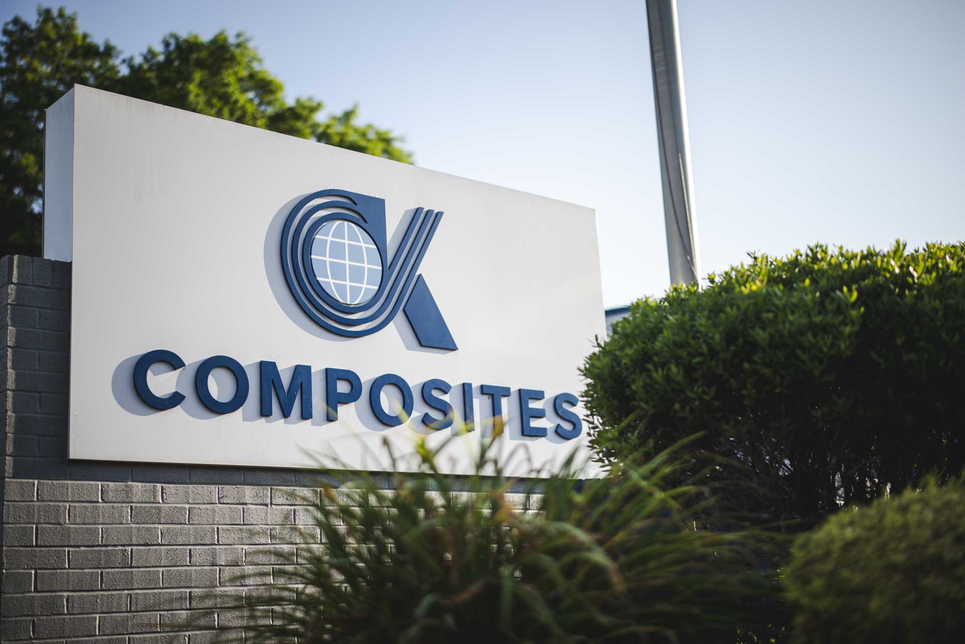 Large outdoor sign with the text OK COMPOSITES and a globe logo stands near green bushes and trees, with a blue sky in the background.