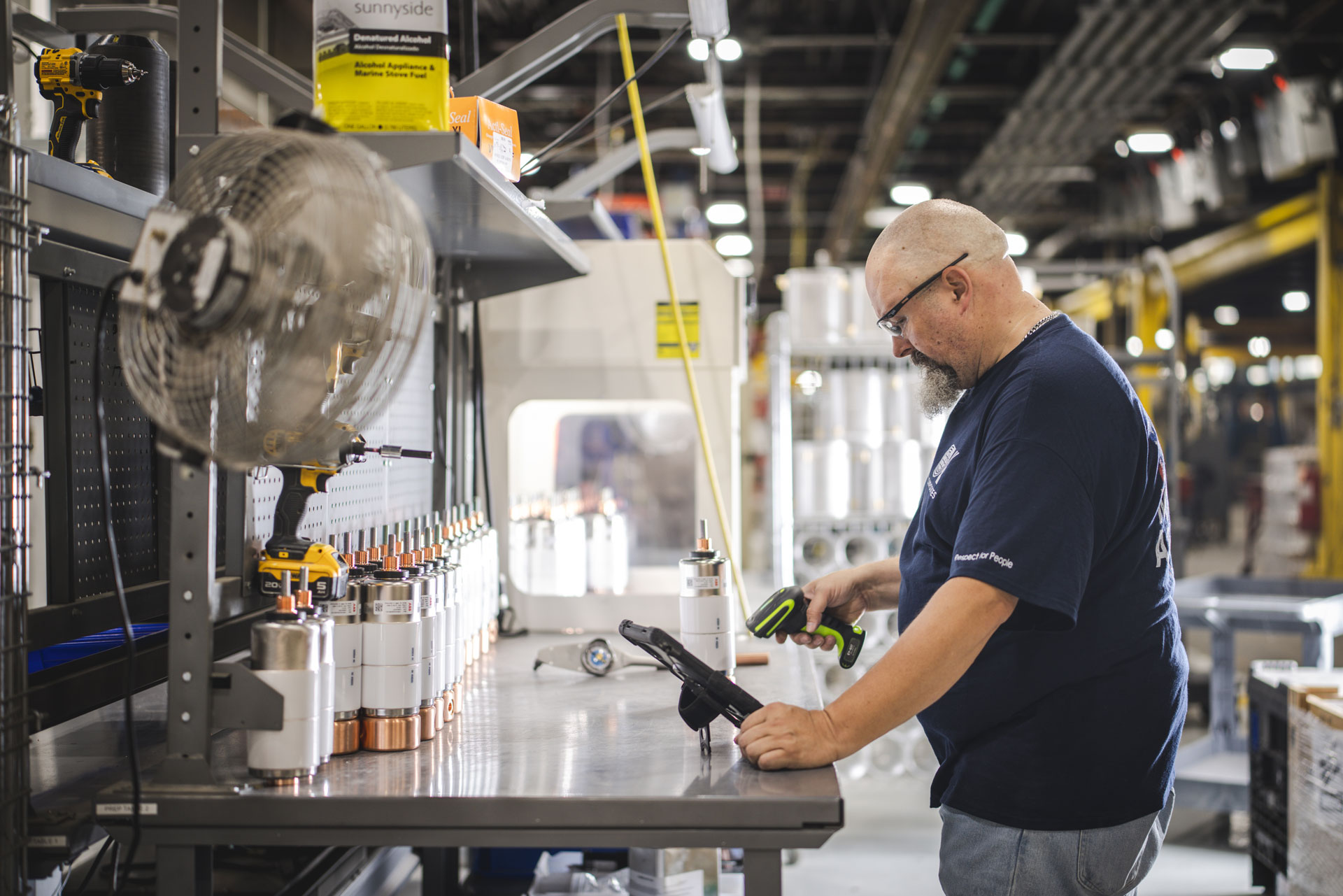 A man in safety glasses uses a power tool at a workbench in a factory, assembling parts. Shelves, tools, and equipment are visible around him, and industrial lighting illuminates the workspace.