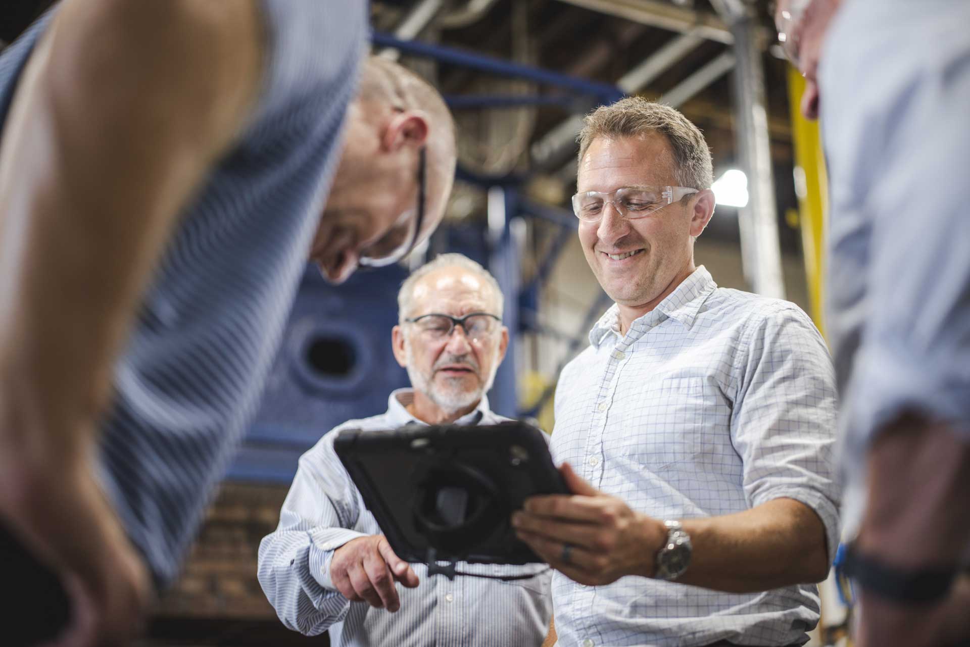 Four men wearing safety glasses stand together in an industrial setting, looking at a tablet and smiling. They appear to be collaborating or reviewing information during a work meeting.