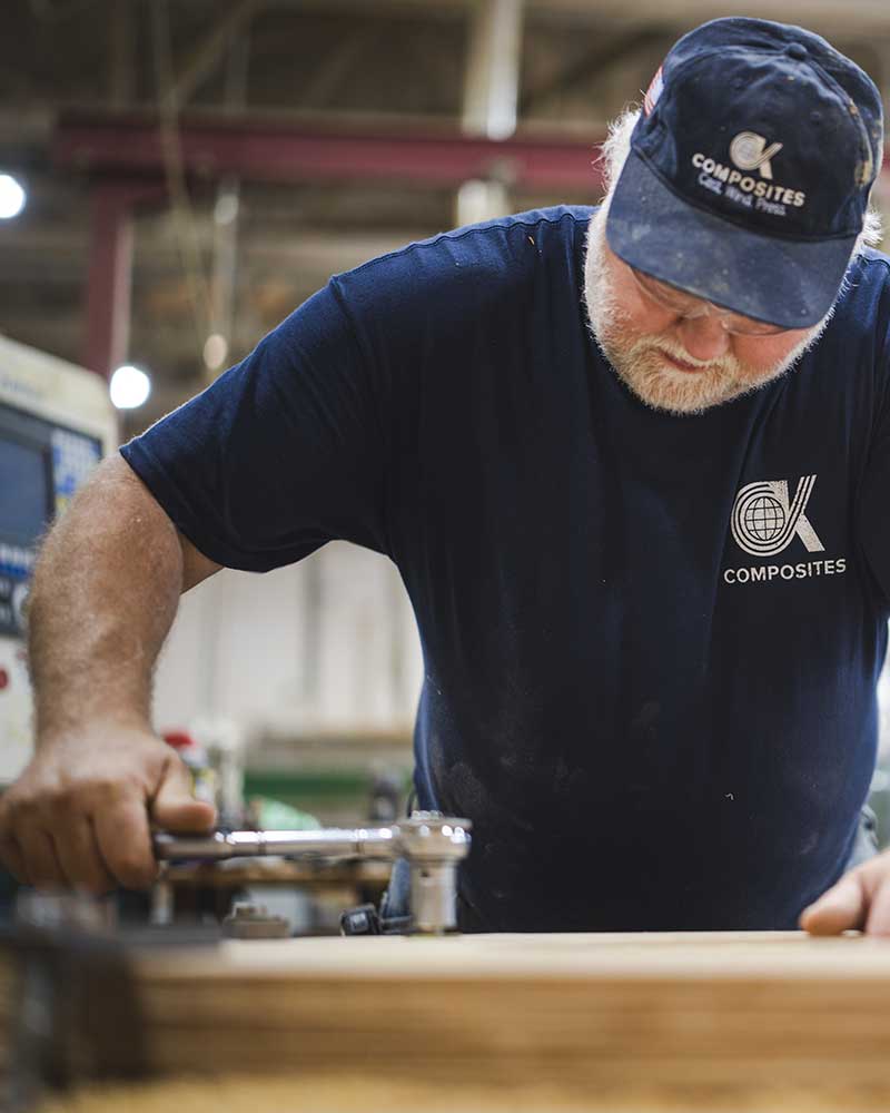 A man in a navy blue shirt and cap works intently with woodworking tools in a workshop, focusing on a piece of wood in front of him.