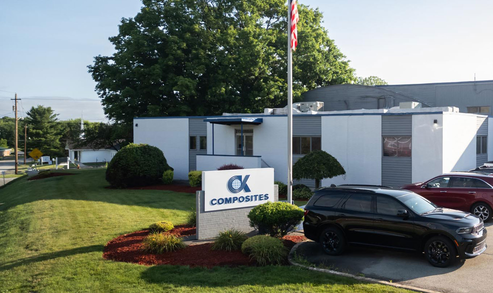 A white building with a sign reading OK Composites stands behind a lawn with bushes and a parked black SUV, with trees and other cars nearby.